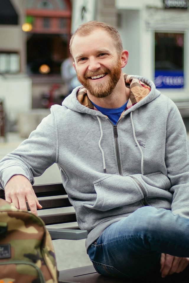 Dating profile photo example: man smiling on a street bench, shot by ProfilePics.ca